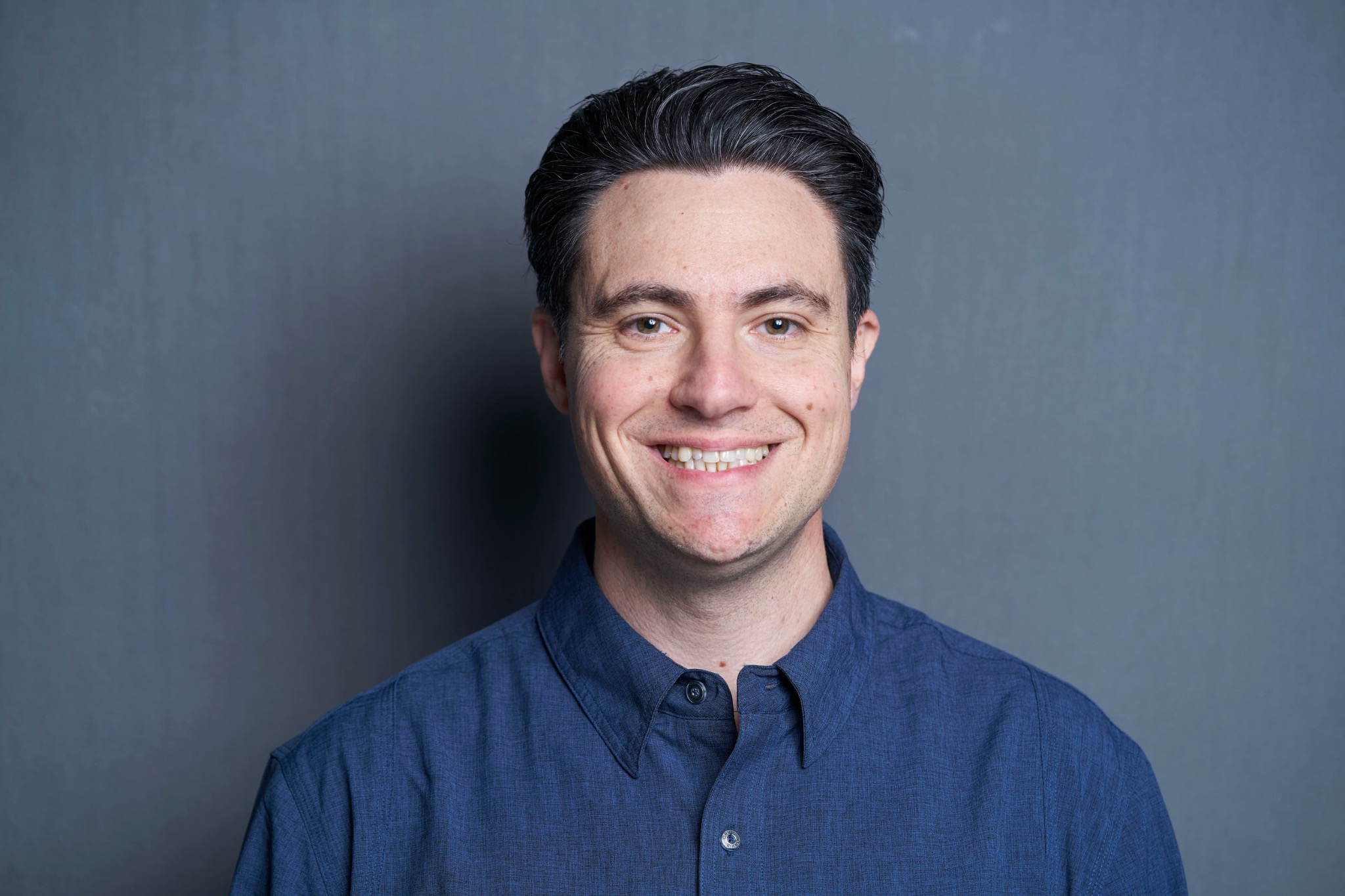 Headshot of Phil Coppola smiling in a blue shirt against a dark background